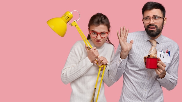 Free Photo pretty woman makes grimace, looks angrily through spectacles, carries table lamp, confused bearded man gestures and looks in displeasure aside