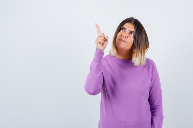 Pretty woman in purple sweater pointing at upper left corner and looking focused , front view.