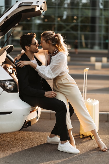 Pretty woman in white outfit smiles widely and hugs boyfriend