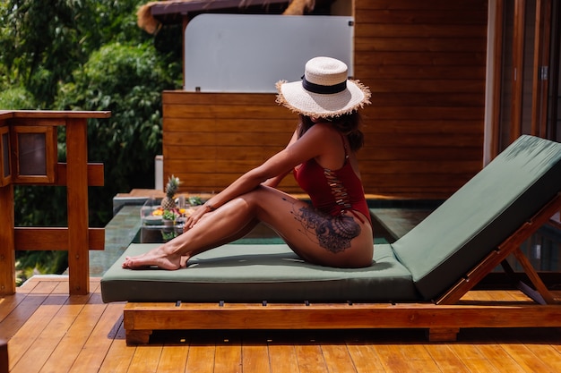 Free photo pretty woman with fit perfect tanned body bronze skin lies on green sunbed at luxury tropical villa in one piece swimsuit with straw hat cover face.