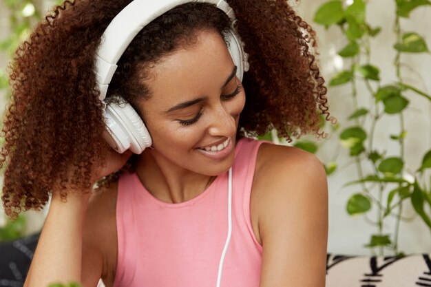 Pretty woman with frizzy dark hair, wears white modern headphones, listens audio track, connected to unrecognizable device. Happy female student listens favourite songs at home, being meloman