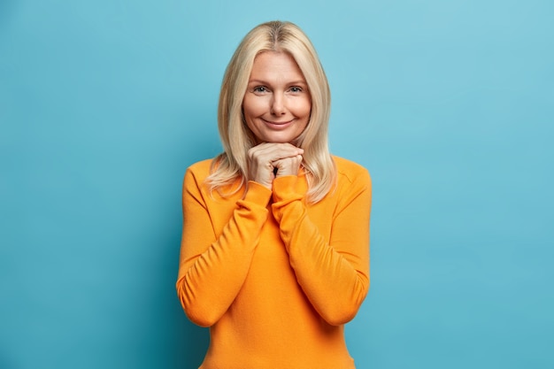 Free photo pretty wrinkled woman with blonde hair keeps hands under chin and looks directly at camera