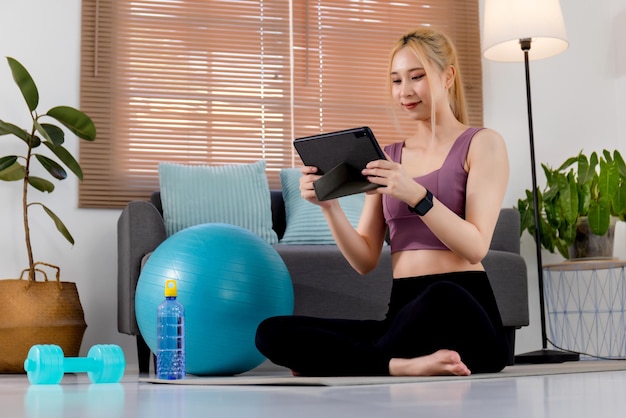 Pretty young Asian woman in sportswear sitting on exercise mat and watching training online videos on digital tablet