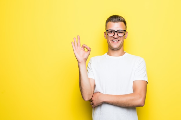 Free photo pretty young boy shows okay sign dressed up in white t-shirt and transperent glasses isolated on yellow