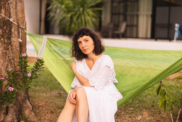 Pretty young caucasian brunette girl wearing white sundress looks at camera sitting on hammock in garden during day