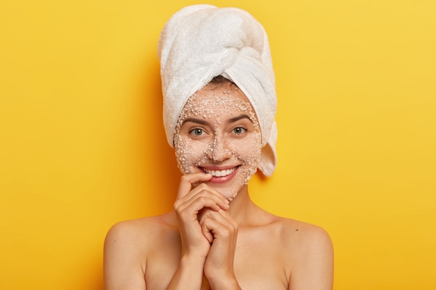 Free photo pretty young european woman enjoys beauty treatments, smiles gently, keeps finger near lips, looks, applies sea salt scrub for peeling complexion