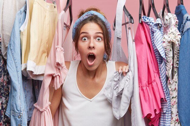 Pretty young female looking with bated breath, posing near hangers with clothes, being shocked to realize that her favourite dress is dirty. People, emotions, body language concept