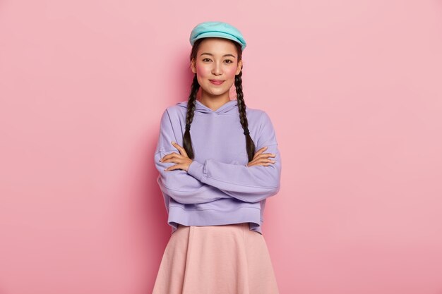 Pretty young female model with satisfied expression, keeps arms folded over chest, looks positively at camera, wears stylish cap, jumper and skirt, has two pigtails, isolated over pink studio wall