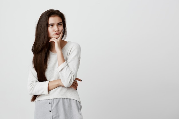 Pretty young female with long dark hair looking away holding hand under chin, building plans, thinking about something. Pensive brunette beautiful woman with thoughtful and pensive face expression