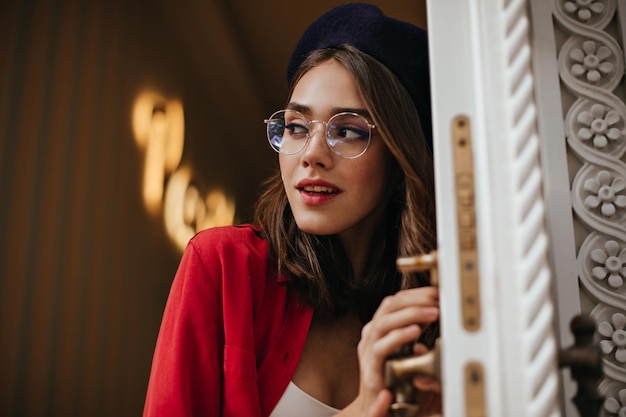 Pretty young girl with brunette hair stylish makeup accessories wearing beret white and red clothes holding door handle and looking away