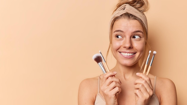 Pretty young lady looks away gladfully uses cosmetic brushes puts on blusher applies decorative makeup on face wears headband casual t shirt stands with bare shoulders isolated over beige background