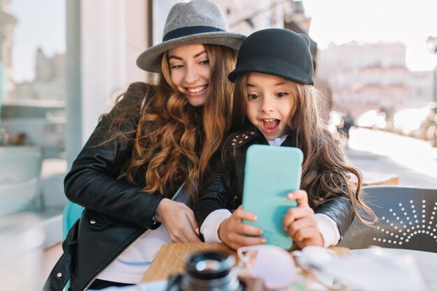 Pretty young mother and her cute daughter having fun and  take selfies. Little girl surprised looking in phone and smile on the sunny city background. Stylish family, true emotion, good mood.