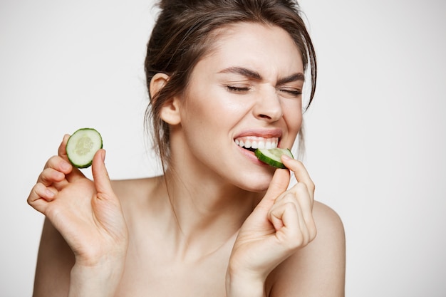 Free photo pretty young natural nude girl with perfect clean skin eating cucumber slice over white background. facial treatment.