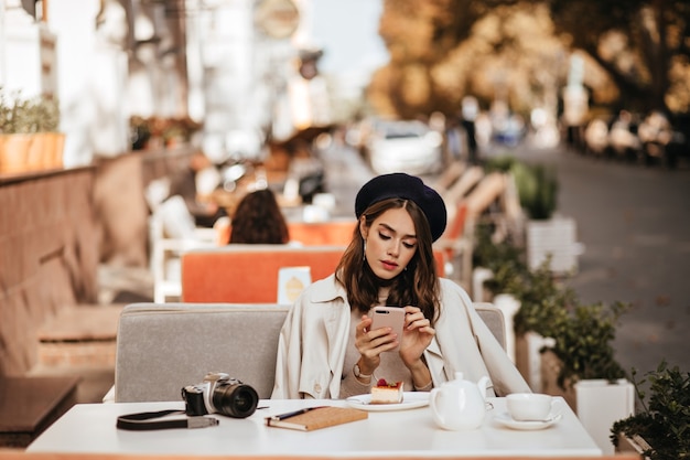 Free Photo pretty young photographer with brunette wavy hairstyle, beret, beige trench coat sitting at city cafe terrace, having tea and cheesecake, holding and looking into mobile phone