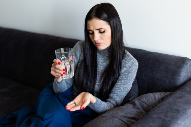 Pretty young sick woman holding a morning after pill and a glass of water at home