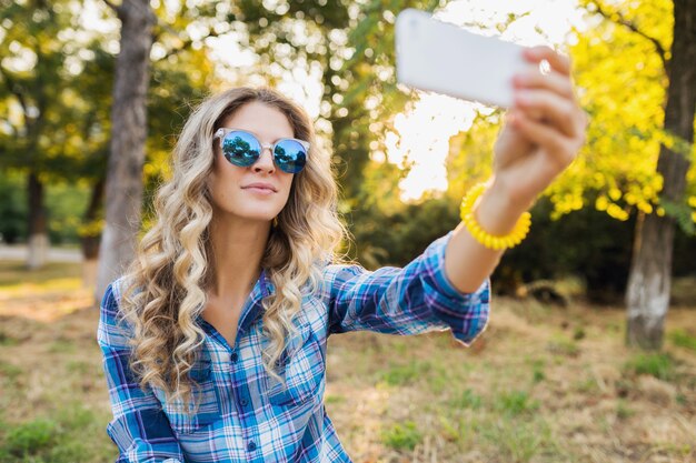 Pretty young stylish attractive smiling blond woman sitting in park, summer casual style