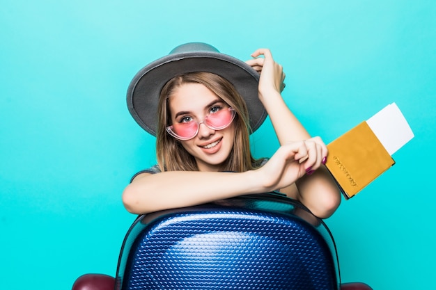 Pretty young teenage lady holds her passport documents with ticket in her hands and blue suitcase isolated on green studio wall