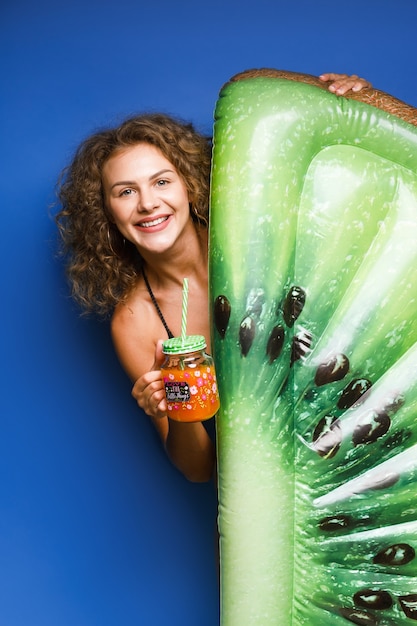 Free photo pretty young woman dressed in swimsuit posing with float kiwi and cocktail on blue