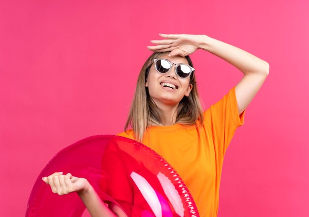 A pretty young woman in an orange t-shirt wearing sunglasses smiling and looking far away while holding pink inflatable ring on a pink wall