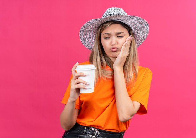 A pretty young woman in an orange t-shirt wearing sunhat keeping hand on teeth while holding plastic cup on a pink wall