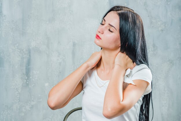 Pretty young woman suffering from neck pain sitting against gray wall