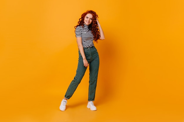 Pretty young woman touches her curly red hair and smiles. Portrait of girl in jeans and top in orange studio.