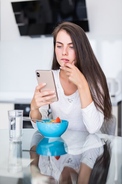 Pretty young woman using her mobile phone while eating salad in the kitchen at home.