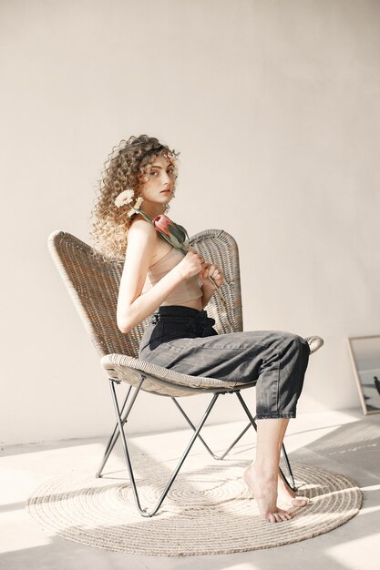 Pretty young woman with flowers has a photosession. Girl with curly hair sitting on a chair.