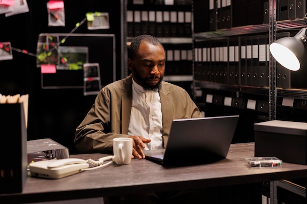 Free photo private investigator sitting at desk and working on laptop in agency at night. concentrated african american criminalist searching crime case file on computer in police detective office