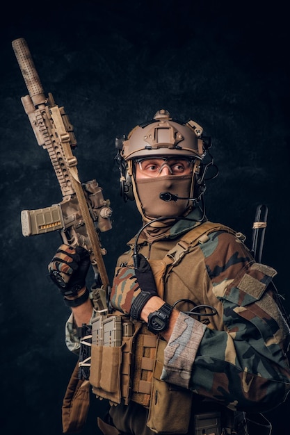 Private security service contractor in camouflage uniform posing with assault rifle. Studio photo against a dark textured wall