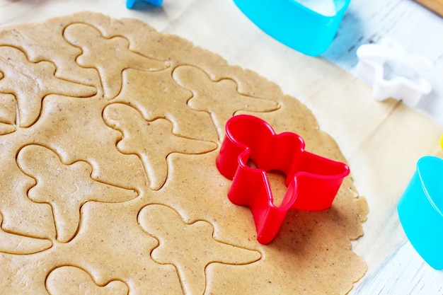 Free photo process of dealing with gingerbread man cookies, use red gingerbread man mold cutting gingerbread dough on baking paper around colorful cookie cutters on white wooden table. top view