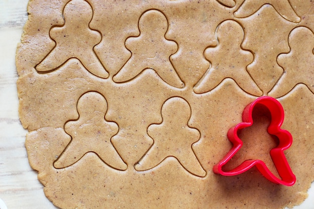 Free photo process of dealing with gingerbread man cookies, use red gingerbread man mold cutting gingerbread dough on baking paper around colorful cookie cutters on white wooden table. top view