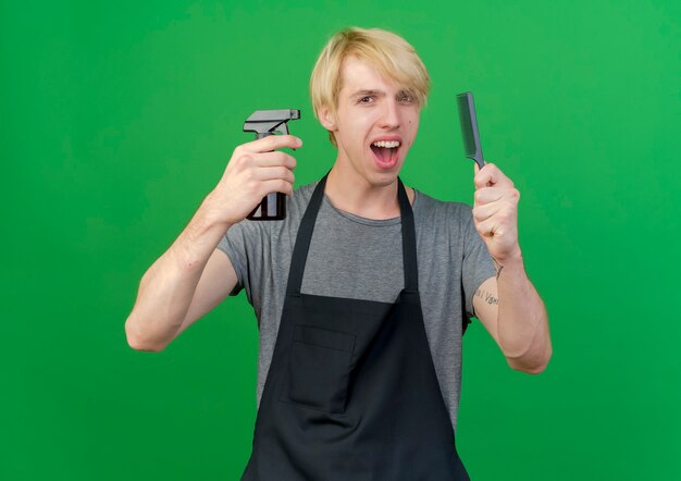 Professional barber man in apron holding hair comb and spray looking happy and positive smiling standing over green background