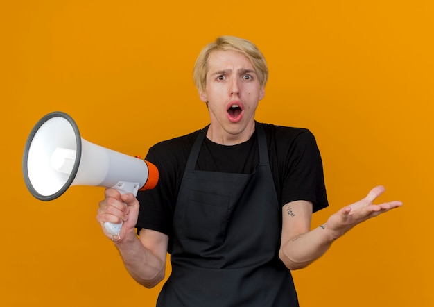 Professional barber man in apron holding megaphone looking at front being surprised and confused standing over orange wall