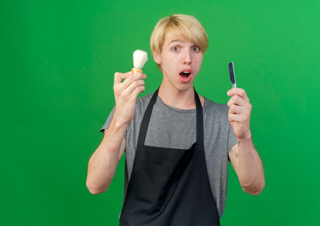 Professional barber man in apron holding razor and shaving brush surprised 