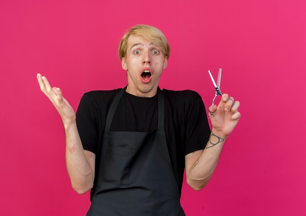 Professional barber man in apron holding scissors being confused and surprised raising hand 