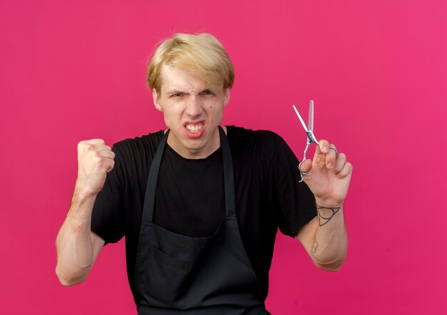 Professional barber man in apron holding scissors clenching fist with angry face 