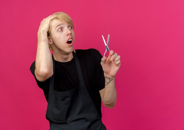 Professional barber man in apron holding scissors lookign aside with hand on his head being confused and frustrated 