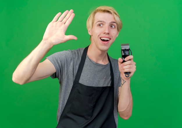 Professional barber man in apron holding trimmer waving with hand smiling friendly standing over green wall