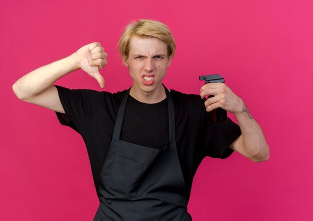 Professional barber man in apron holding trimmer with angry face showing thumbs down 