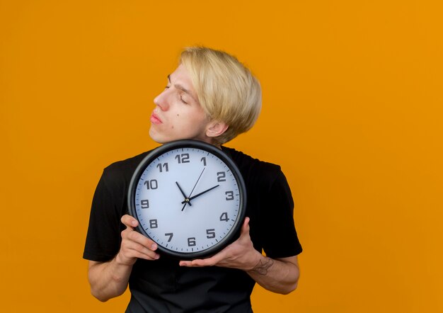 Professional barber man in apron holding wall clock looking aside with serious face standing over orange wall