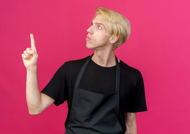 Professional barber man in apron looking aside confused pointing with index finger at something 