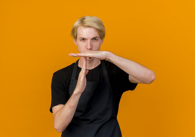 Free photo professional barber man in apron looking at front with serious face making time out gesture with hands standing over orange wall