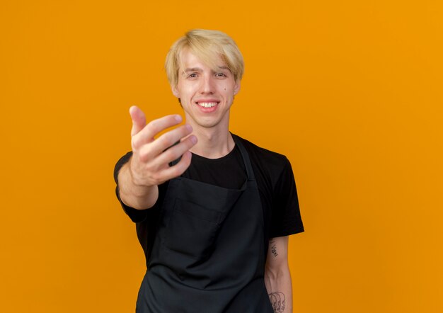 Professional barber man in apron making come here gesture with hand smiling friendly standing over orange wall