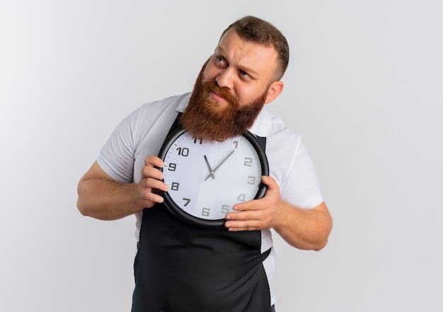 Professional bearded barber man in apron holding wall clock looking aside very anxious standing over white wall