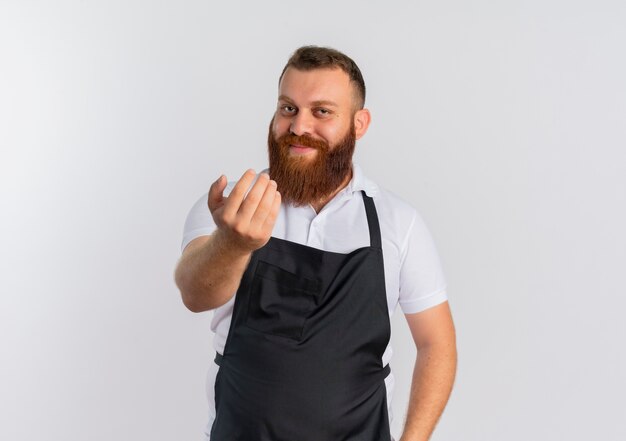 Professional bearded barber man in apron smiling friendly inviting to come standing over white wall