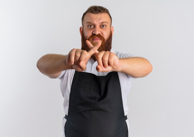 Professional bearded barber man in apron with angry face making stop sign crossing index fingers standing over white wall