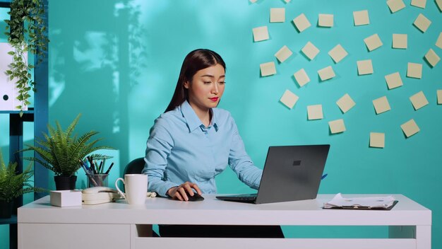 Professional businesswoman bored in administration office, playing with pen while working on laptop. Fatigued employee woman working in modern workplace over blue studio background