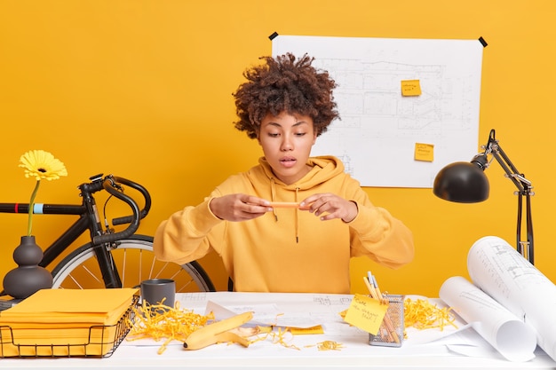 Professional dark skinned young female architect dressed in sweatshirt makes photo of her sketches on mobile phone sits at desktop 