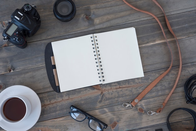 Free photo professional digital camera; cup of coffee; blank spiral notepad; pen; spectacles and cable on wooden table